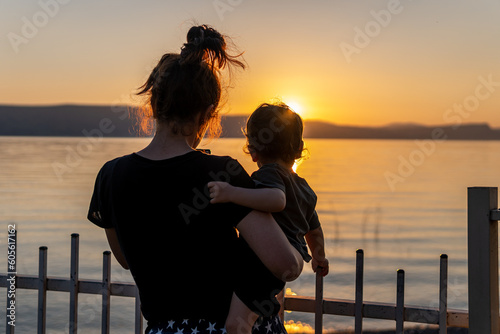 Silhouettes of a mother holding her child in her arms looking at the beautiful sunset and lake. Yellow and orange sky with sun rays and mom enjoying with her young infant out in nature. Family time.