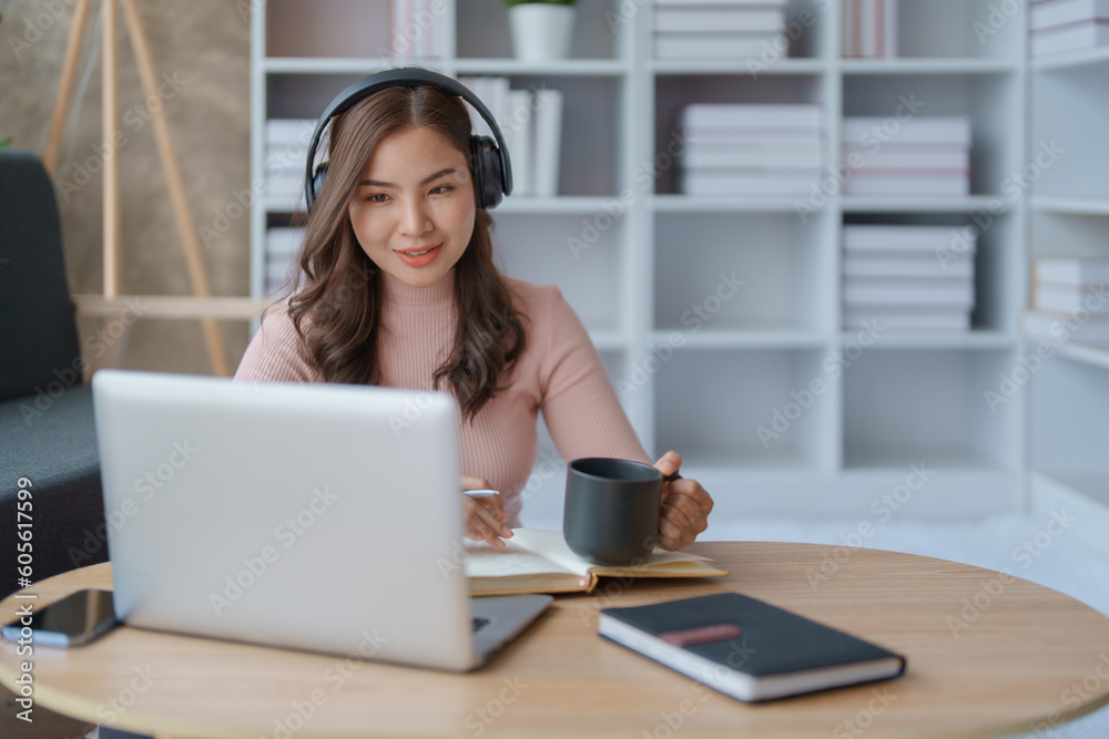 Portrait of a teenage Asian woman using a laptop computer, wearing headphones and using a notebook to study online via video conferencing and holding coffee cup on a wooden desk in living room. Portrait of a teenage Asian woman using a laptop computer, wearing headphones and using a notebook to study online via video conferencing and holding coffee cup on a wooden desk in living room.
