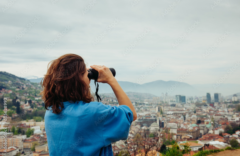 A young adult female traveler looks through binoculars at a European city with orange roofs in the mountains