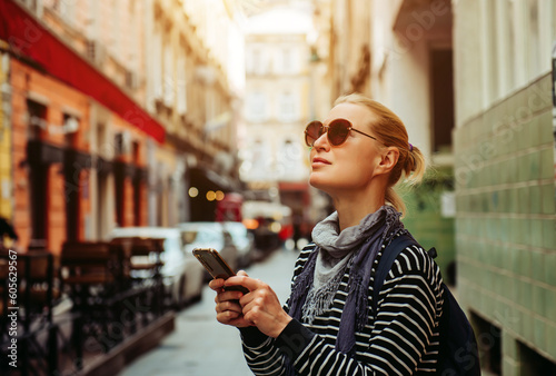A young adult blonde female traveler walks and smiles around a European city, using a smartphone, taking photos and studying information from travel apps