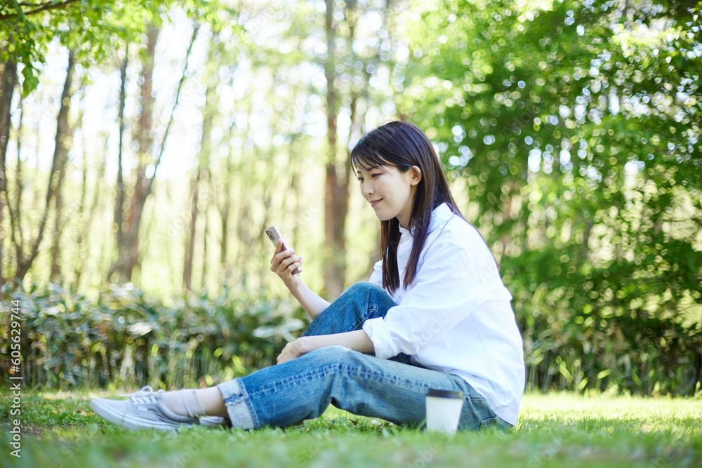 森の中でリラックスしながらスマートフォンを操作する30代日本人女性の初夏のイメージ 森の中でリラックスしながらスマートフォンを操作する30代日本人女性の初夏のイメージ