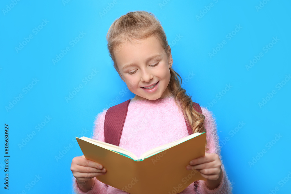 Happy little girl with backpack reading book on light blue background Happy little girl with backpack reading book on light blue background