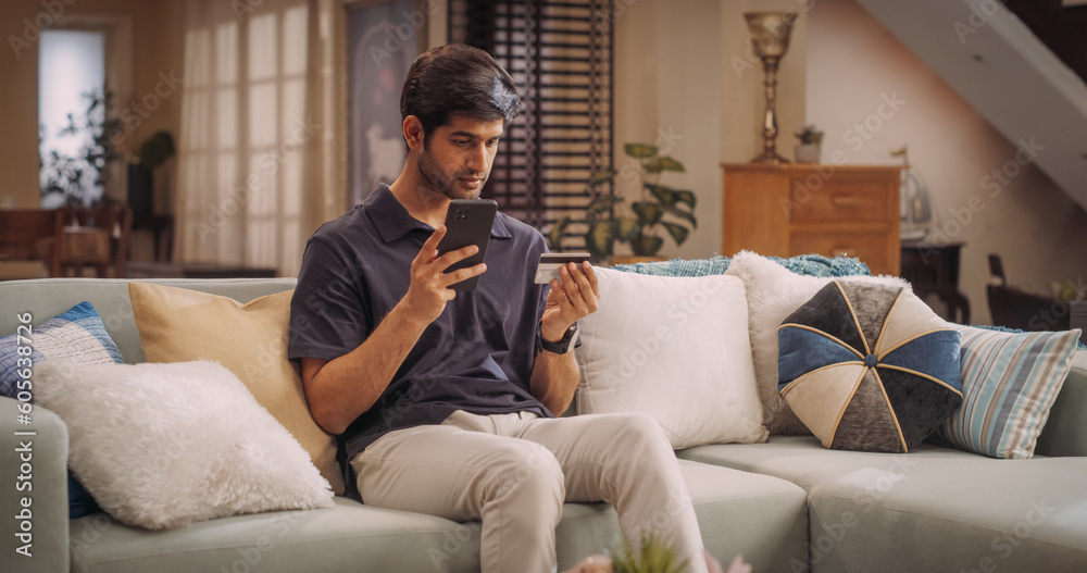 Obraz premium Happy Handsome Indian Man Using Credit Card and Smartphone for Online Shopping at Home. Smiling Happy Guy Purchasing a Surprise Gift for His Girlfriend. e-Commerse Concept. Medium Shot 