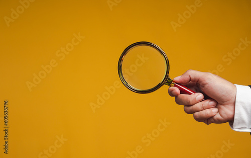Close-up of a businessman's hand holding a magnifying glass while standing on an orange background