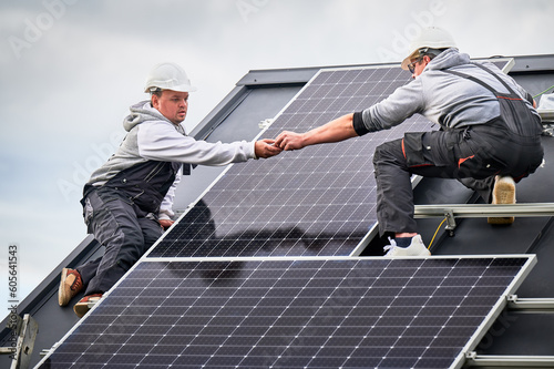 Workers building photovoltaic solar module station on roof of house. Men electricians in helmets installing solar panel system outdoors. Concept of alternative and renewable energy.