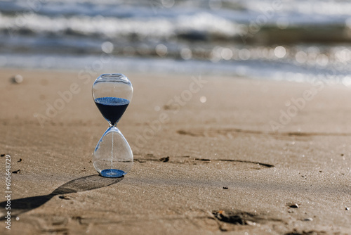 Hourglass with blue sand inside in a warm golden morning sunlight with shadow on a sandy beach and sea background, starting time for a new day or running of time. Concept of the rapid passage of time photo