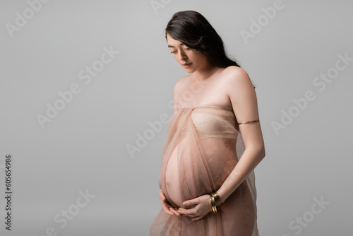elegant expecting mother with wavy brunette hair, standing in beige chiffon cloth and golden bracelets isolated on grey background, maternity fashion concept, pregnant woman