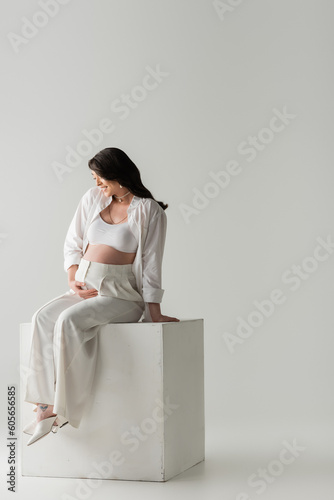 full length of smiling mother-to-be in stylish pants, crop top and shirt touching tummy while sitting on white cube on grey background in studio, maternity fashion concept, pregnant woman