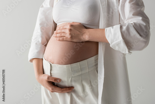 partial view of future mother in white stylish shirt, crop top and pants tenderly embracing tummy isolated on grey background, maternity fashion concept, pregnant woman