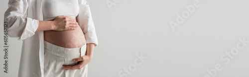 partial view of future mother in white crop top, shirt and pants embracing belly while standing isolated on grey background, maternity fashion concept, banner, pregnant woman