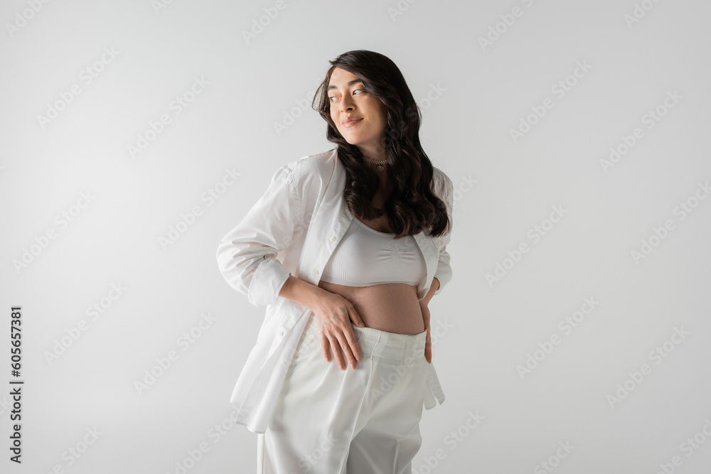 cheerful pregnant woman in white stylish shirt, crop top and pants touching tummy and looking away isolated on grey background, maternity fashion concept