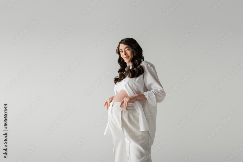 happy future mother with wavy brunette hair posing in white fashionable clothes such as white shirt and pants isolated on grey background, trendy maternity concept happy future mother with wavy brunette hair posing in white fashionable clothes such as white shirt and pants isolated on grey background, trendy maternity concept