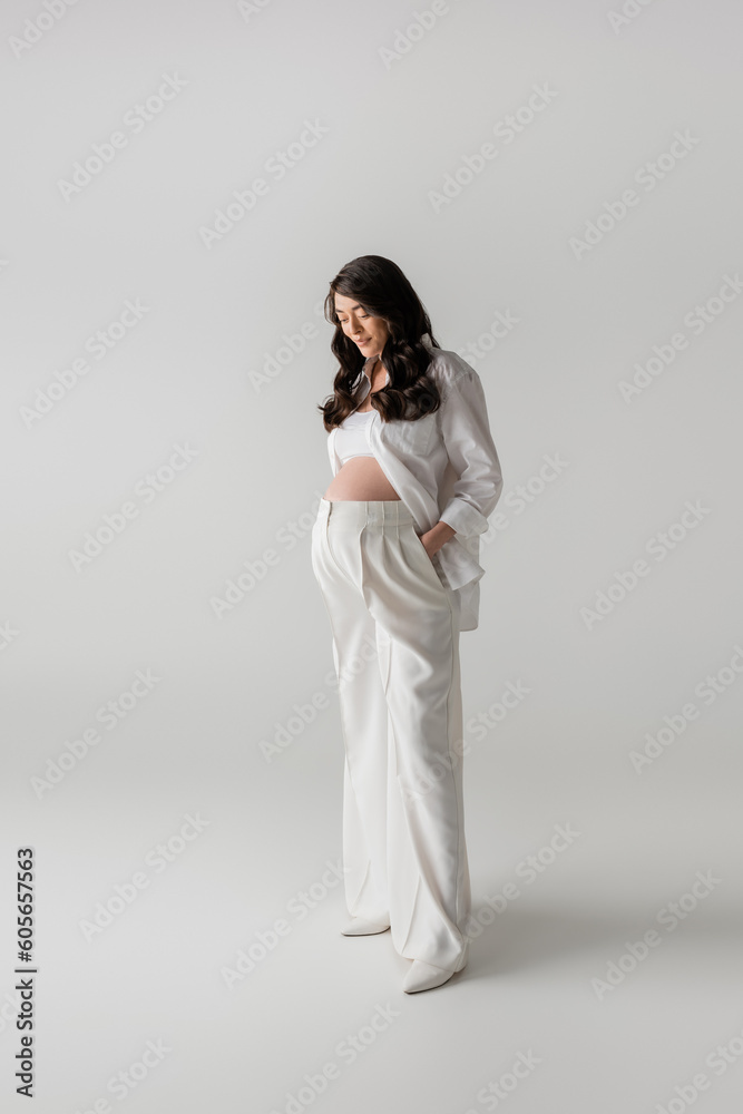 full length of brunette pregnant woman in white shirt and crop top standing with hand in pocket of pants on grey background, fashionable pregnancy concept, expectation, future mother with belly full length of brunette pregnant woman in white shirt and crop top standing with hand in pocket of pants on grey background, fashionable pregnancy concept, expectation, future mother with belly