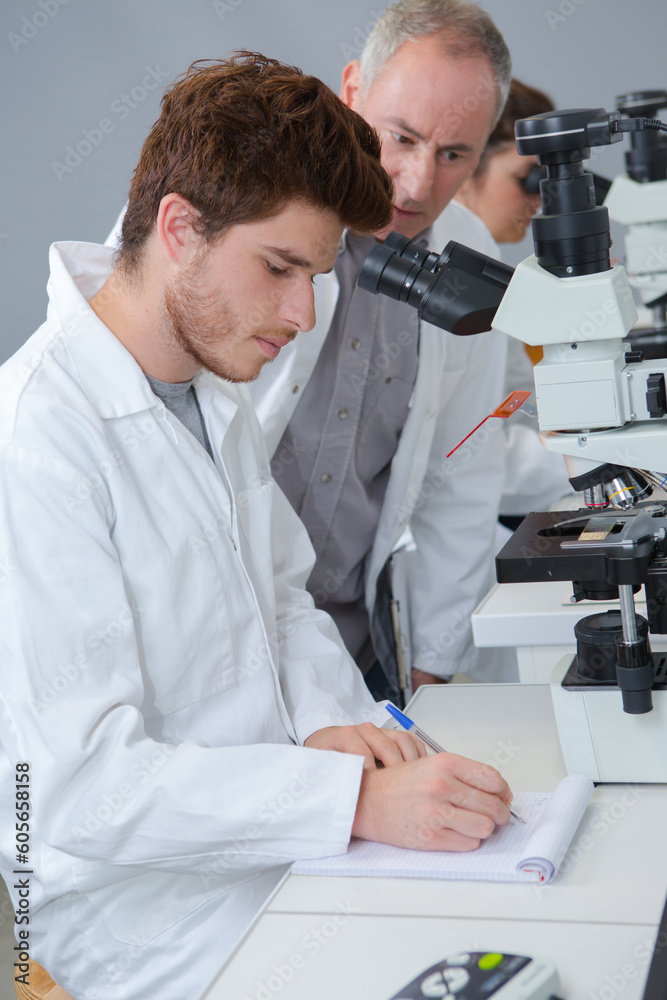 male biology teacher looking through microscope in classroom - obrazy, fototapety, plakaty 