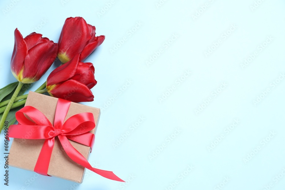 Beautiful gift box with bow and red tulips on light blue background, flat lay. Space for text