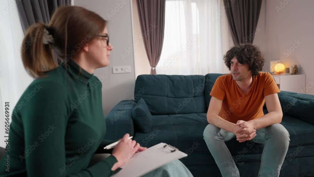 unhappy American man sitting on couch in counseling office and complaining about his troubles to female psychologist while she taking notes on clipboard unhappy American man sitting on couch in counseling office and complaining about his troubles to female psychologist while she taking notes on clipboard