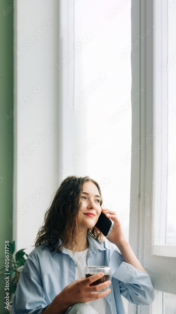 Cheerful young woman in casual attire chatting and flirting with her boyfriend on the phone, sitting on a windowsill with a cup of black tea.
