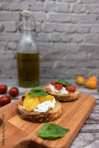 Tasty bruschetta with ricotta cheese, red and yellow cherry tomatoes and basil on a wooden cutting board. Ingredients on the background, vertical image with bokeh effect.