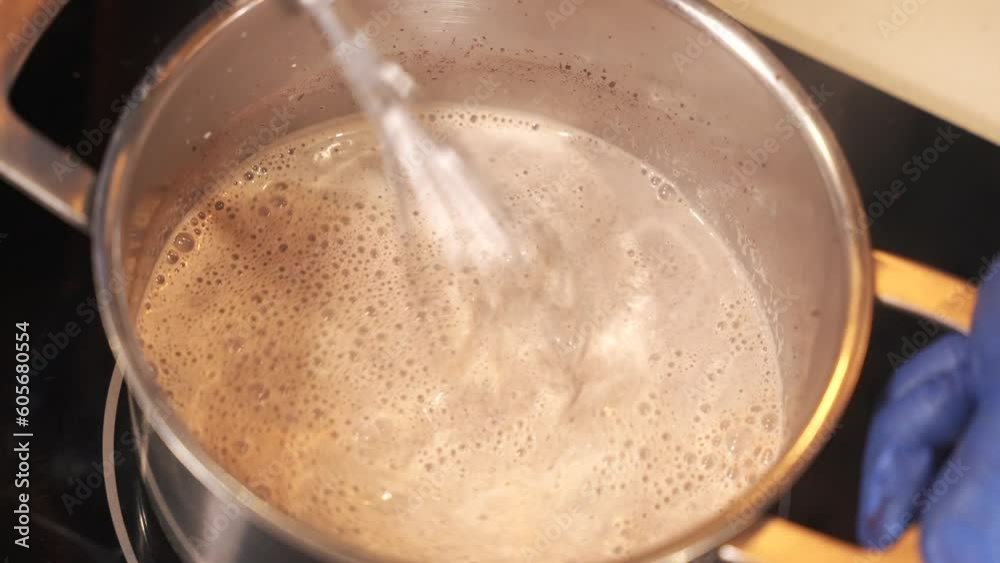 Close up of a chef hands in gloves whisking hot chocolate with the milk in a stainless steel pot on the electric stove in the kitchen. Cooking delicious drink Close up of a chef hands in gloves whisking hot chocolate with the milk in a stainless steel pot on the electric stove in the kitchen. Cooking delicious drink