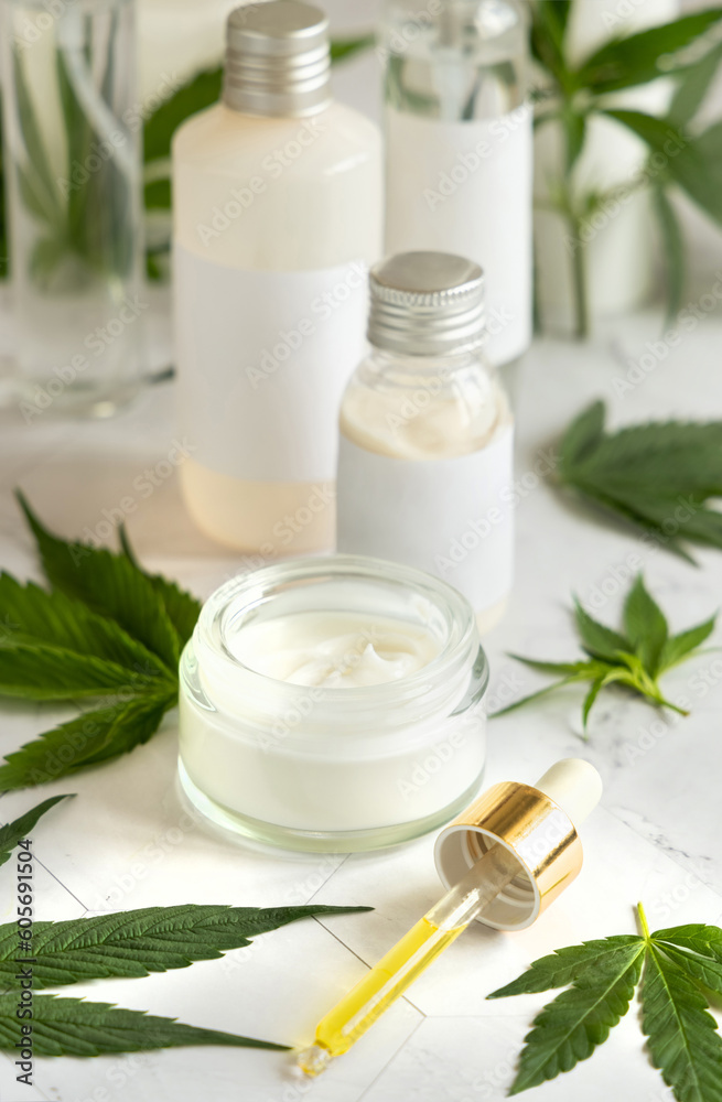 Blank Cream jar and bottles near green cannabis leaves on white table. Cosmetic Mockup