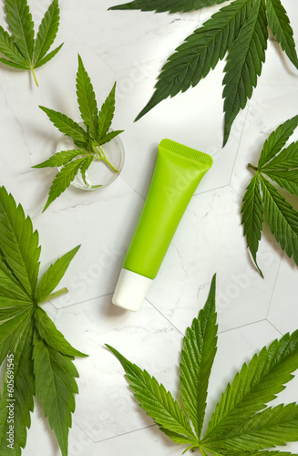Green Blank Cream tube near green cannabis leaves on white table top view. Cosmetic Mockup