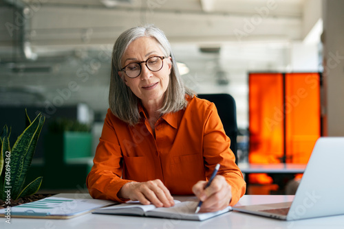 Confident mature woman, writer  using laptop computer, taking notes, working project in modern office. 60 years old female, financier working at workplace. Successful business