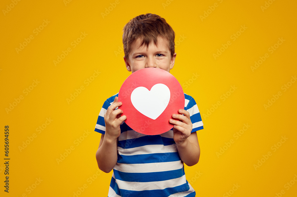 Delighted little kid in blue striped shirt, keeping heart icon near face while representing social media for kids against yellow background Delighted little kid in blue striped shirt, keeping heart icon near face while representing social media for kids against yellow background