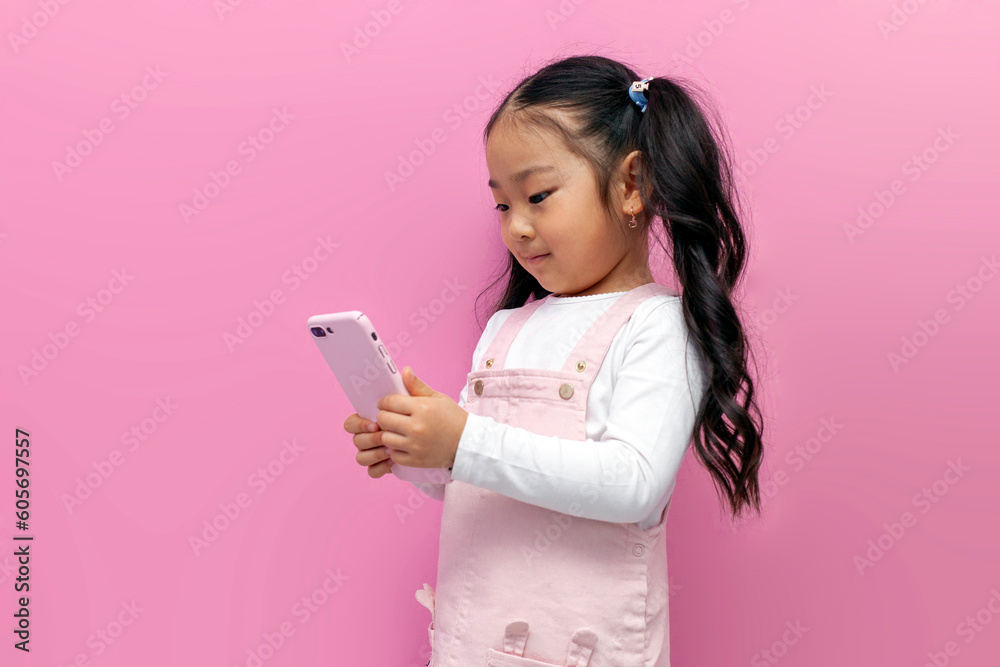 little asian girl in pink sundress uses smartphone and chooses online on pink isolated background