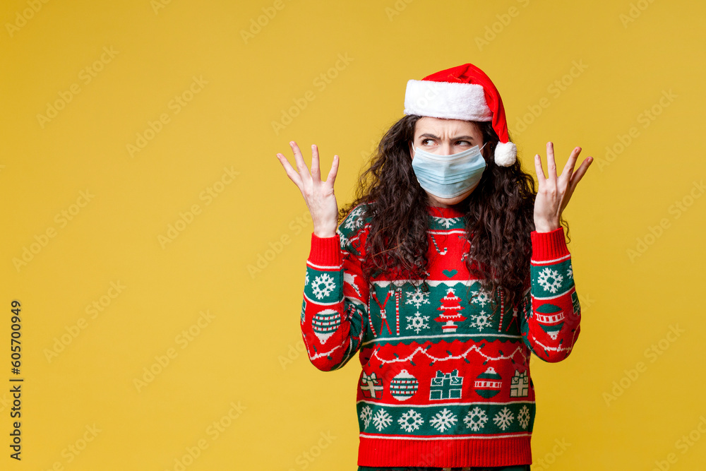 young perplexed girl in christmas clothes and santa hat in medical protective mask shows bewilderment