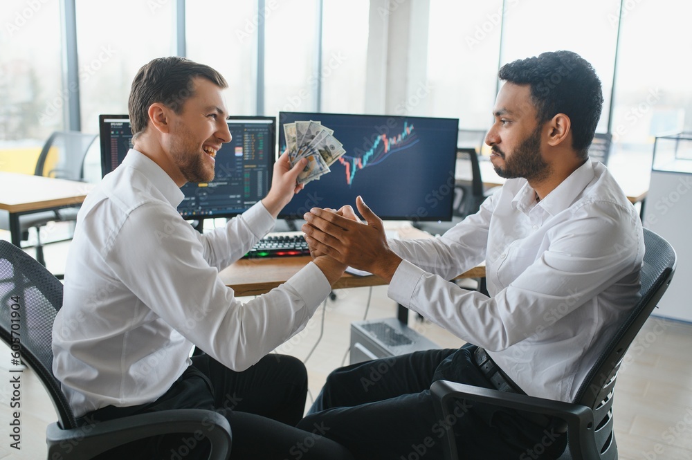 Obraz premium Two men traders sitting at desk at office together monitoring stocks data candle charts on screen analyzing price flow smiling cheerful having profit teamwork concept