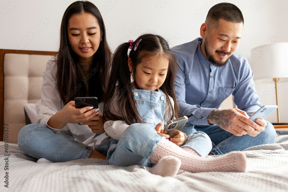 happy young asian family use smartphones at home on bed, korean dad mom and little daughter look at phone