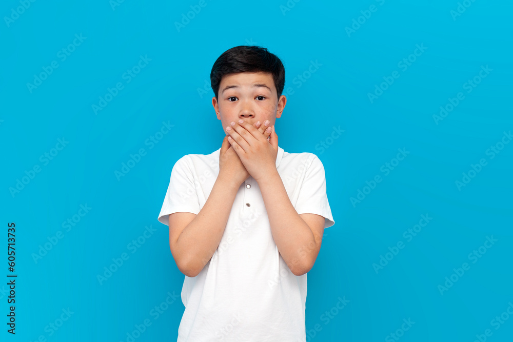 shocked little asian boy in white covers his mouth with his hand on blue background, korean child is silent shocked little asian boy in white covers his mouth with his hand on blue background, korean child is silent