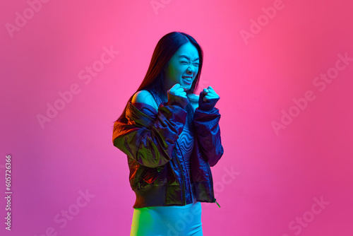 Anger, irritation. Portrait of young korean girl in casual clothes posing with fists up against pink studio background in neon light. Concept of emotions, facial expression, youth, lifestyle, ad