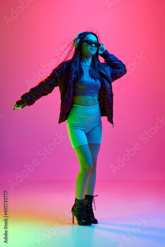 Portrait of pretty, young girl in sunglasses, listening to music in headphones against pink studio background in neon light. Concept of emotions, facial expression, youth, lifestyle, inspiration, ad