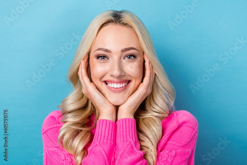 Photo of adorable gorgeous girl with wavy hairdo dressed pink sweater palms on cheekbones toothy smiling isolated on blue color background © deagreez