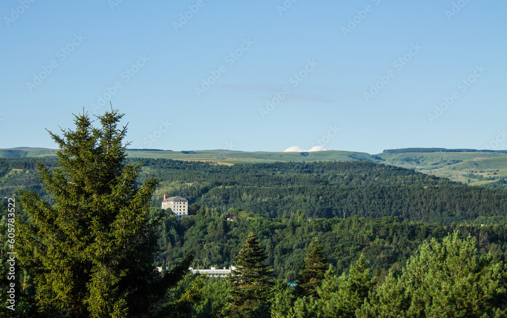 Panoramic view of the picturesque green hilly valley and the snowy peak of Mount Elbrus blurred in a misty haze on a clear sunny summer day Panoramic view of the picturesque green hilly valley and the snowy peak of Mount Elbrus blurred in a misty haze on a clear sunny summer day
