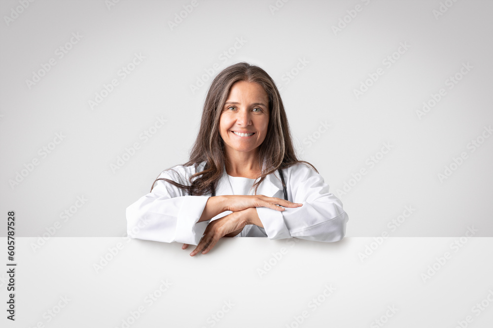 Smiling confident senior woman doctor in white coat with stethoscope near big banner with empty space on gray background Smiling confident senior woman doctor in white coat with stethoscope near big banner with empty space on gray background
