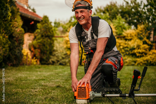 Senior man with brush cutter on the lawn against the background of bushes in workwear and goggles