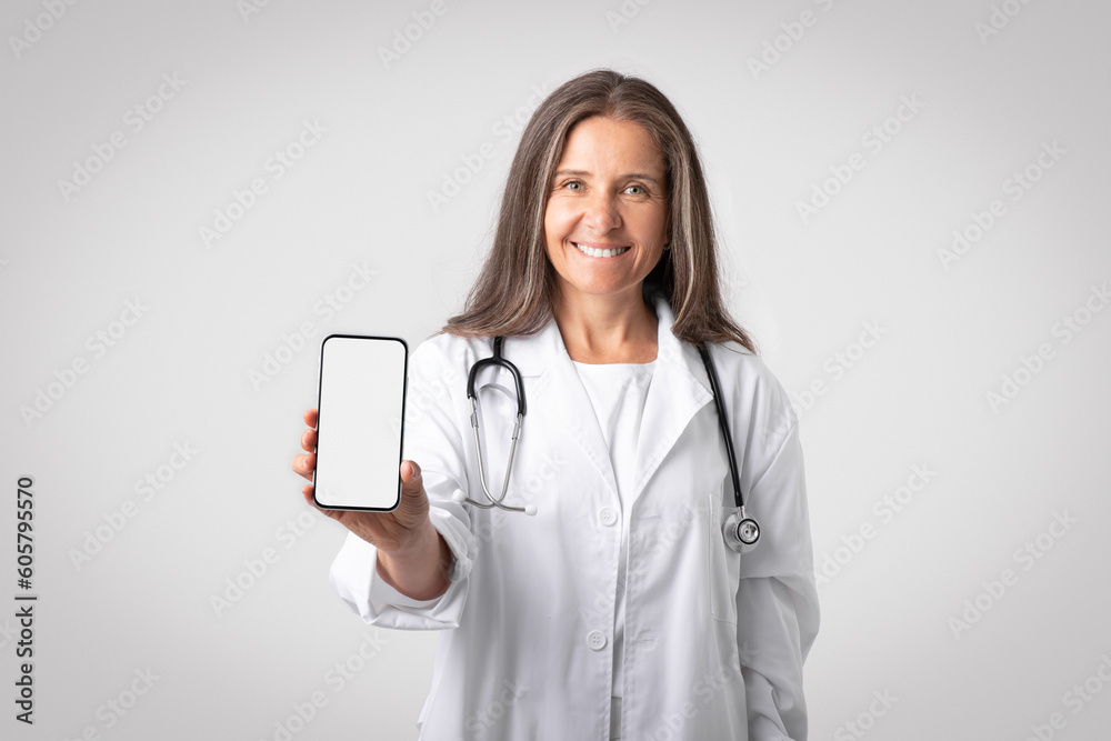 Friendly senior woman doctor in white coat with stethoscope showing phone with white blank screen to camera, mockup Friendly senior woman doctor in white coat with stethoscope showing phone with white blank screen to camera, mockup