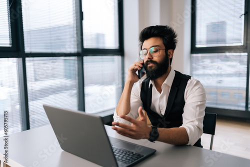 Successful Indian bearded business man in glasses working at laptop computer sitting at desk and talking on smartphone by window. Handsome businessman in elegant formalwear talking to client