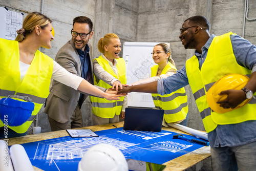 Team of structural engineers and architects holding hands together above building plans and blueprints at construction site.