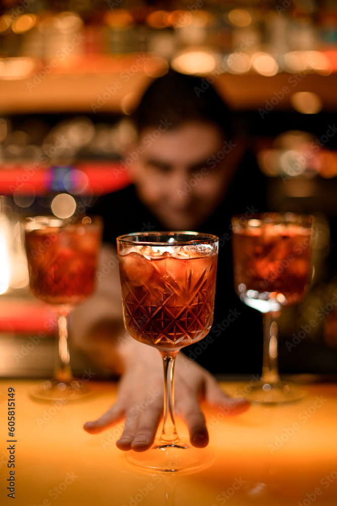 Male bartender presents cold red cocktail with ice cubes in glass on the bar counter in the bar Male bartender presents cold red cocktail with ice cubes in glass on the bar counter in the bar