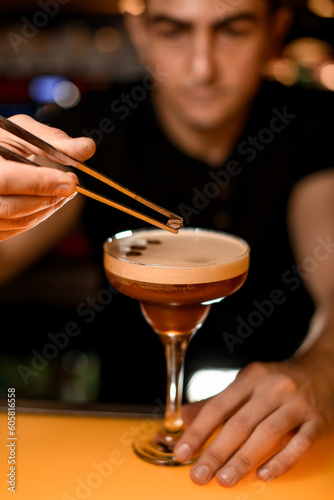 Male bartender's hand neatly holds glass with cold cocktail and garnishes it with coffee bean using tweezers