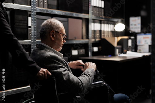 Paralyzed private detective wheelchair user and assisting coworker in criminal cases archive room. Senior investigator with disability surrounded by criminology folders on cabinet shelves