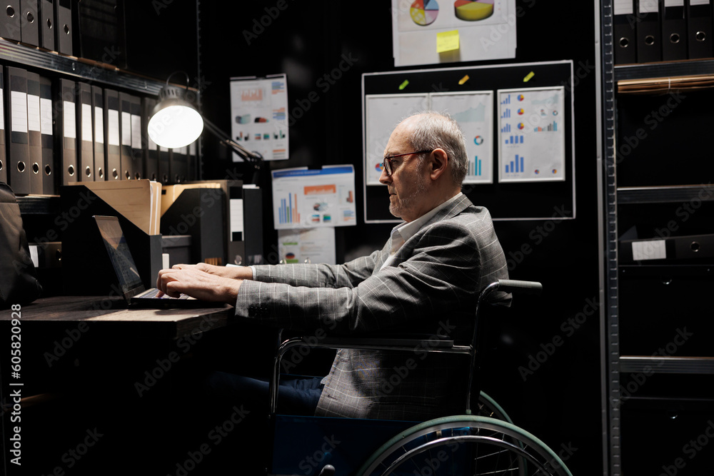 Senior businessman employee with disability in wheelchair inserting accountancy budget plan data on laptop. Bureaucratic executive in storage room office filled with chart reports and document folders - obrazy, fototapety, plakaty 