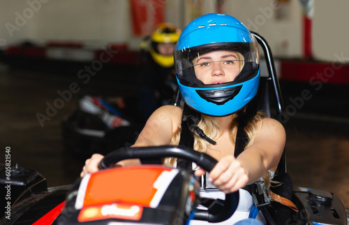serious woman in helmet driving car for karting with other people in sport club indoor