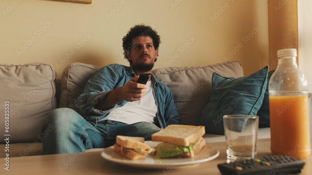 Guy dressed in denim shirt has lunch when he sits in the living room with remote control in his hands
