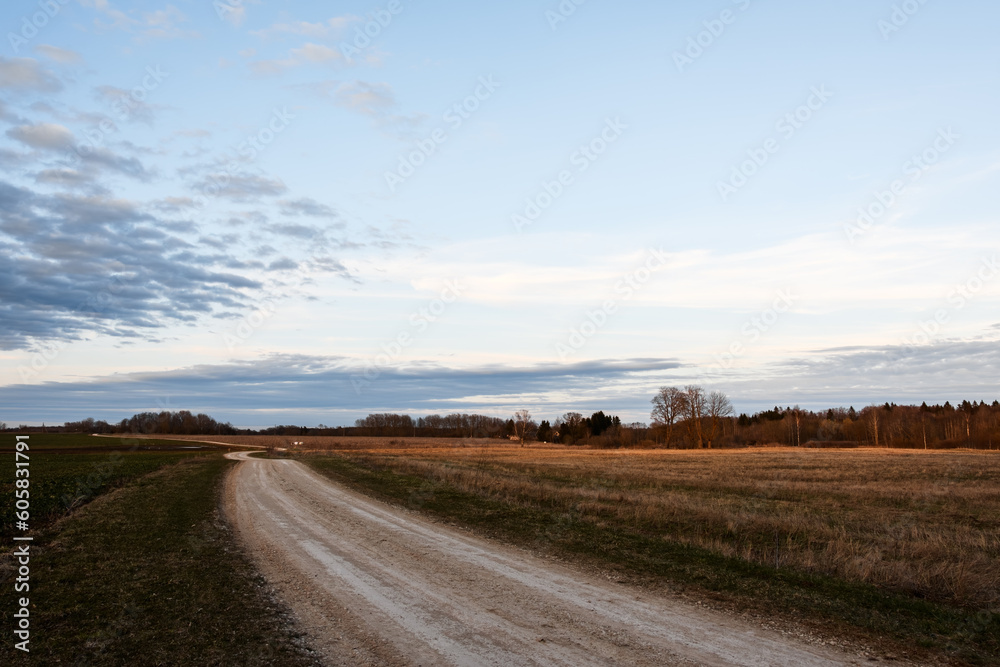 Winding dirt country road. Landscape of agricultural fields in spring. Evening blue sky with gray and white clouds. A twilight shadow falls to the ground