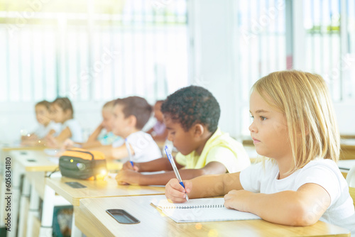 Focused cute towheaded preteen schoolgirl writing exercises in workbook in classroom during lesson