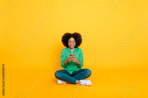 Full-length photo of happy positive trendy african american or brazilian curly woman, sits on a yellow background, uses her smartphone, chats on social networks, browsing internet, news, smiles